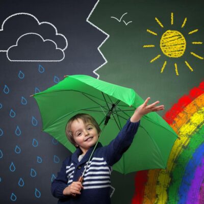Child holding an umbrella standing in front of a chalk drawing of changing weather from rain storm to sun shine with a rainbow on a school blackboard